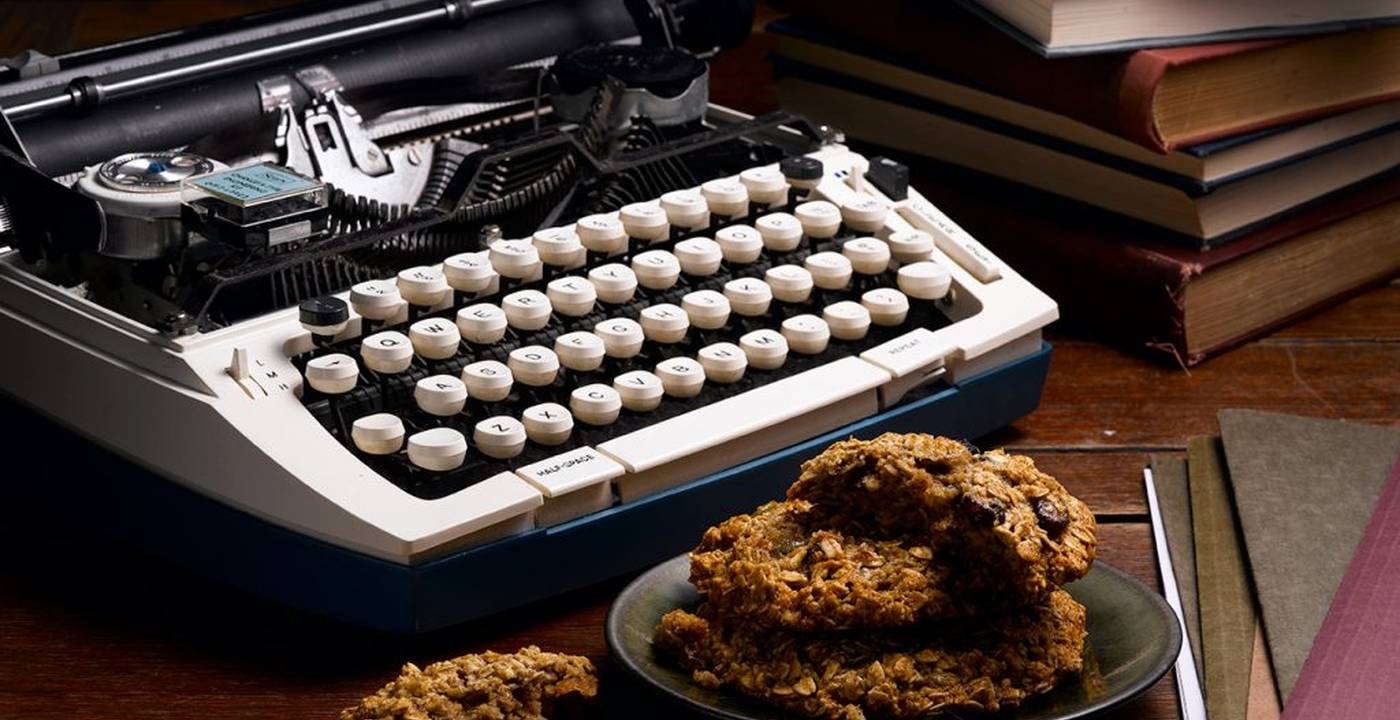 A vintage typewriter on a wooden desk beside a plate of oat cookies and a stack of old books.